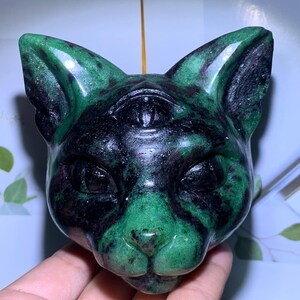 May include: A carved stone cat head sculpture featuring a deep green and black mottled pattern. The feline figure has pointed ears and a prominent third eye carved into the center of the forehead, with smooth, polished surfaces and defined facial features.