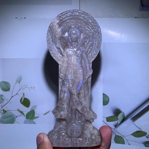 以下が含まれることがあります： 頭の後ろに円形の光背を背負った立像の神を描いた、半透明で淡い紫色の彫刻クリスタルフィギュア。彫刻は衣服や台座に複雑なディテールが施されており、磨き上げられた滑らかで半透明な質感が特徴です。
