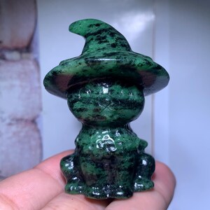 May include: A small, carved stone figurine of a cat wearing a pointed witch hat. The sculpture is made of a dark green mineral with black speckled inclusions throughout the surface, featuring a smooth, polished texture and a seated posture.