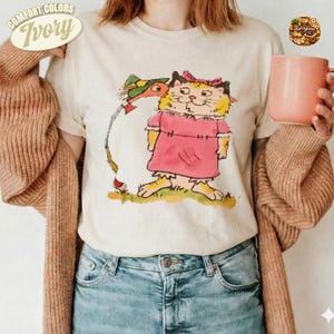 Könnte beinhalten: Ein cremefarbenes T-Shirt mit einer grafischen Illustration einer gelben Katze in einem rosa Kleid und grünem Hut, kombiniert mit einer braunen Strickjacke und blauen Jeans. Ein einfarbiger rosa Becher wird seitlich gehalten. Text COMFORT COLORS IVORY.