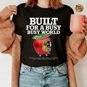Könnte beinhalten: Ein schwarzes T-Shirt mit dem weißen Text BUILT FOR A BUSY BUSY WORLD über einer Grafik eines roten Apfelautos mit einem grünen Blatt oben und einer Wurmfigur darin. Unter dem Auto steht der Text RICHARD SCARRY'S BUSY WORLD FEATURING LOWLY WORM AND HIS APPLE CAR.
