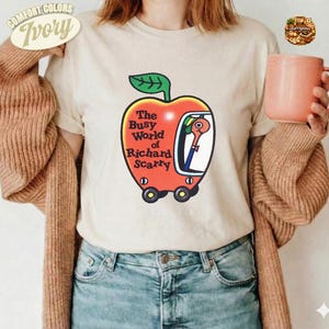 Könnte beinhalten: Ein elfenbeinfarbenes T-Shirt mit einer Grafik eines roten Apfels in Form eines Autos mit einem grünen Blatt oben. Im Inneren des Apfelautos befindet sich ein Wurm. Der Text lautet The Busy World of Richard Scarry. Getragen mit einer braunen Strickjacke und blauen Jeans.