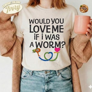 Könnte beinhalten: Ein elfenbeinfarbenes T-Shirt mit dem schwarzen Text "WOULD YOU LOVE ME IF I WAS A WORM?" über einer Grafik eines Zeichentrickwurms mit grünem Hut und rosa Herz. Das Shirt wird unter einer beigen Strickjacke getragen, kombiniert mit blauer Jeans.