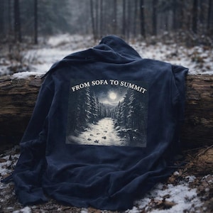 Pu&ograve; includere: Una felpa con cappuccio blu navy con una grafica di una scena di foresta sul retro, raffigurante un sentiero che conduce a una luna piena. Il testo "FROM SOFA TO SUMMIT" &egrave; sopra la grafica. La felpa &egrave; drappeggiata su un tronco in una foresta innevata.