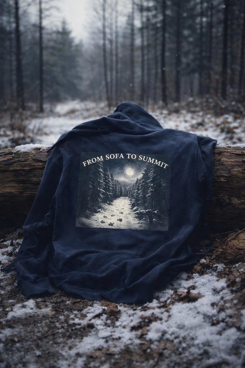 Pu&ograve; includere: Una felpa con cappuccio blu navy con una grafica di una scena di foresta sul retro, raffigurante un sentiero che conduce a una luna piena. Il testo "FROM SOFA TO SUMMIT" &egrave; sopra la grafica. La felpa &egrave; drappeggiata su un tronco in una foresta innevata.