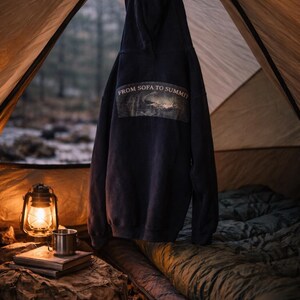 Pu&ograve; includere: Una felpa blu navy con la scritta "FROM SOFA TO SUMMIT" &egrave; appesa all'interno di una tenda. Una lanterna accesa, una tazza di metallo e uno zaino sono su dei libri. Un sacco a pelo &egrave; visibile.