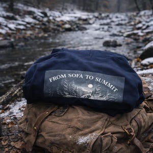 Pu&ograve; includere: Una felpa blu navy con la scritta "FROM SOFA TO SUMMIT" stampata sul retro, appoggiata su uno zaino marrone. Una tazza da campeggio in metallo &egrave; posizionata vicino. La scena &egrave; all'aperto con un ruscello innevato sullo sfondo.