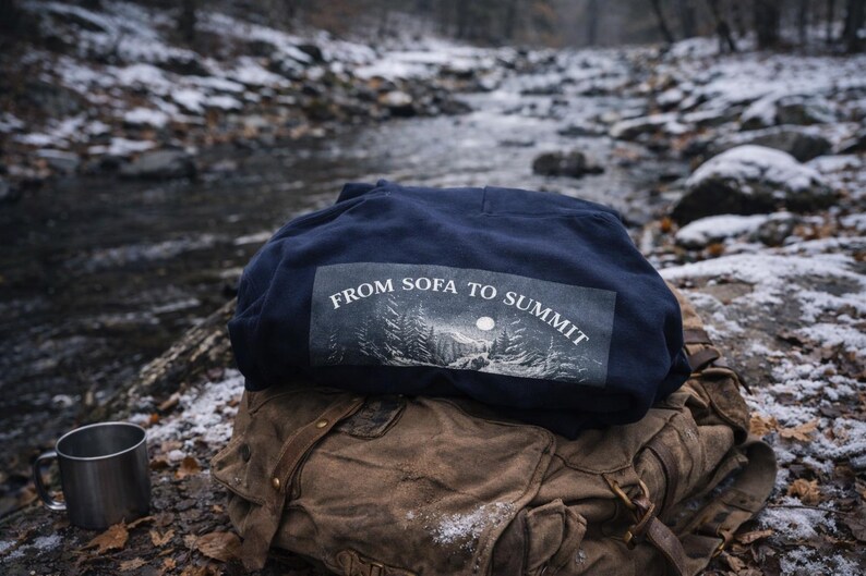 Pu&ograve; includere: Una felpa blu navy con la scritta "FROM SOFA TO SUMMIT" stampata sul retro, appoggiata su uno zaino marrone. Una tazza da campeggio in metallo &egrave; posizionata vicino. La scena &egrave; all'aperto con un ruscello innevato sullo sfondo.