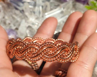 Könnte beinhalten: Ein kupferfarbenes Metallarmband mit einem durchgehenden, komplizierten geflochtenen Drahtdesign. Das Armband besteht aus mehreren Strängen gedrehtem Draht, die zu einem sich wiederholenden wellenförmigen Muster gewebt sind.