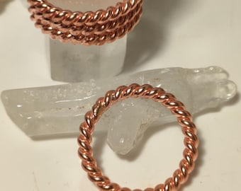 Könnte beinhalten: Kupferfarbene Ringe aus verdrilltem Draht. Ein Ring wird separat gezeigt, während drei auf einem klaren Kristall gestapelt sind. Die Ringe haben ein geflochtenes Aussehen und ein glänzendes Finish. Es sind Schmuckstücke.