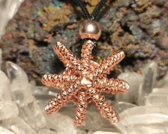 Könnte beinhalten: Ein Anhänger aus Kupferdraht in Form eines mehrarmigen Sterns oder Meerestiers mit eng gewickelten Drahtsegmenten. Der Anhänger hängt an einer schwarzen Schnur mit einer einzelnen Metallperle über dem Stern, vor einem strukturierten Mineral.