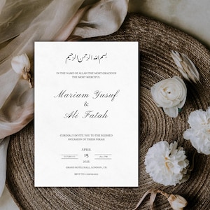 May include: A white wedding invitation with elegant black calligraphy. The card features names, date, and location details, and is adorned with white flowers. It rests on a textured, brown surface. The invitation is for Mariam Yusuf and Ali Fatah.