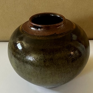 May include: A round ceramic vase with a glossy, mottled dark green and brown glaze. The vessel features a wide, spherical body that tapers toward a short, dark brown neck with a smooth, circular opening at the top for floral arrangements.