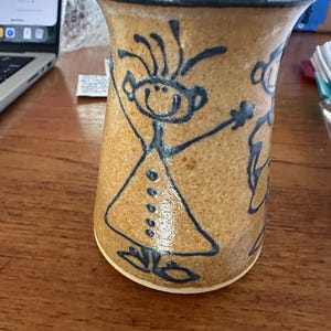 May include: A tan ceramic mug with a dark brown rim and a hand-drawn dark blue line art illustration of a person with spiky hair wearing a dress with buttons. The mug has a flared top opening and a speckled texture throughout the exterior surface.