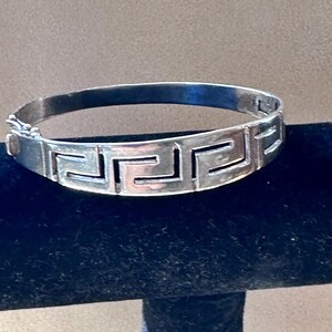 Könnte beinhalten: Ein silberfarbener Klapparmreif mit einem sich wiederholenden geometrischen griechischen Mäandermuster, das in die Außenfläche eingraviert ist. Das Armband verfügt über einen sicheren Metallverschluss an der Seite für eine geschlossene Form.