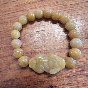 May include: A stretch bracelet composed of smooth, round, pale yellow beads and a single central carved bead shaped like a mythical creature. The beads have a translucent quality and a polished finish, creating a uniform appearance throughout the piece.