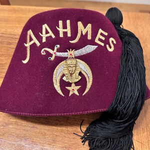 以下が含まれることがあります： 金色の刺繍で「AAHMES」と記されたマルーン色のフェルト製フェズ帽。銀色の刺繍が施されたシミター、金色の金属製ファラオのエンブレム、赤い石があしらわれた五芒星が特徴です。側面には長い黒いタッセルが垂れ下がっています。