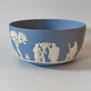 May include: A light blue ceramic bowl featuring white relief decorations of classical figures and trees. The matte finish highlights the raised, detailed carvings of people in draped garments standing among foliage, creating a distinct contrast.