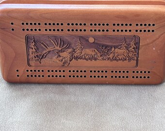 Peut inclure: Un plateau de cribbage rectangulaire en bois avec une finition lisse brun clair. Le plateau présente une scène centrale gravée au laser représentant deux cerfs dans un décor forestier avec des arbres et un soleil. Des rangées de petits trous.