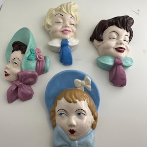 Puede incluir: Cuatro placas de pared de cerámica con forma de rostros estilizados. Una presenta a una persona con un sombrero verde menta y un lazo morado. Otra tiene cabello rubio y una corbata azul. Una tercera muestra cabello oscuro con una corbata verde azulado.