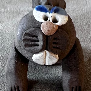 May include: A dark brown clay figurine of a beaver with a textured surface. The figure features large white eyes with blue pupils, a small tan nose, and prominent white front teeth. It has two front paws with etched lines for claws.