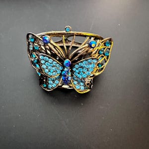 May include: A metal napkin ring featuring a butterfly design with an antique bronze finish. The butterfly wings are embellished with numerous small, bright blue rhinestones and larger, darker blue faceted stones along the center body.