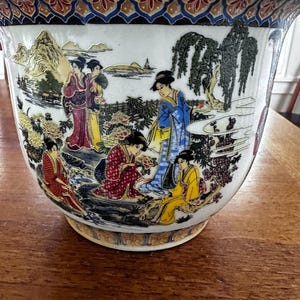 May include: A white ceramic planter featuring a colourful wrap-around illustration of figures in traditional clothing, trees, and mountains. The rim and base are decorated with a repeating geometric pattern in blue, gold, and red.