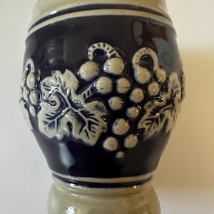May include: A stoneware mug featuring a dark blue glazed body with a contrasting beige rim and base. The exterior is decorated with a raised, embossed relief pattern of grape clusters and vine leaves in a light beige color against the blue background.