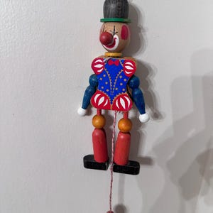 May include: A wooden clown jumping jack toy featuring a black hat, a red nose, and a blue torso with red and white accents. The figure has blue arms with white hands, orange joints, red legs, and black feet, suspended by a red string with a red bead.
