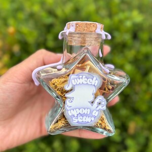 Könnte beinhalten: Eine sternförmige Glasflasche mit Korken und lavendelfarbenen Wachsakzenten. Die Flasche enthält getrocknete Kräuter und einen Aufkleber mit der Aufschrift "witch upon a star". Der Hintergrund ist verschwommenes grünes Laub.