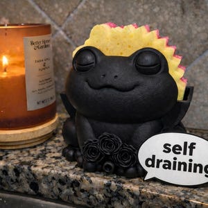 May include: A black frog-shaped sponge holder with a yellow and pink sponge. The holder has a smiling face and a cluster of roses. A speech bubble says "self draining". A lit candle in a glass jar is to the left.