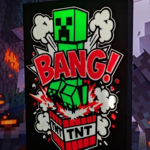 May include: Black rectangular sign featuring a green pixelated character exploding above a red and white TNT box. The word "BANG!" is in large red letters with white accents. White cloud-like shapes surround the explosion.