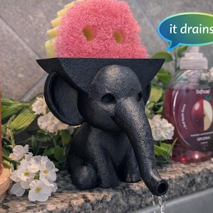 May include: A black elephant-shaped sponge holder with a long trunk spout, holding a pink and yellow sponge. The text "it drains!" is in a speech bubble. The holder sits on a countertop with white flowers and a bottle of soap.