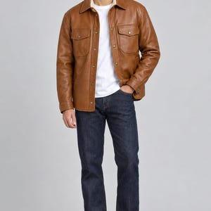 May include: A tan leather jacket with a pointed collar, two chest pockets with flaps, and metal snap buttons. The jacket is worn over a plain white t-shirt and paired with dark blue denim jeans and brown leather boots, creating a layered look.
