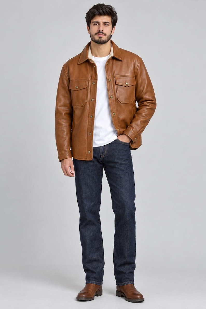 May include: A tan leather jacket with a pointed collar, two chest pockets with flaps, and metal snap buttons. The jacket is worn over a plain white t-shirt and paired with dark blue denim jeans and brown leather boots, creating a layered look.
