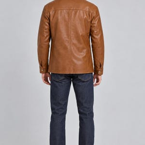 May include: A tan leather jacket with a pointed collar, long sleeves with buttoned cuffs, and vertical seam detailing on the back, paired with dark blue denim jeans and brown leather shoes, viewed from the back against a plain light gray background.