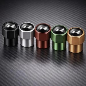 May include: A set of five cylindrical tire valve caps with a hexagonal base, each featuring a black circular top with a white letter R logo. The caps are finished in matte black, silver, copper, green, and gold metallic colors respectively.