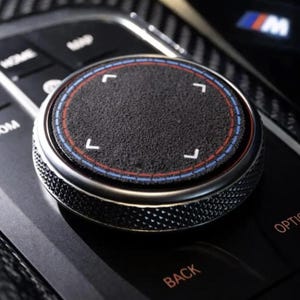 May include: A circular black textured control knob for a vehicle infotainment system, featuring a metallic knurled edge, a central touch-sensitive surface with four white corner markers, and thin red and blue circular accent rings around the perimeter.