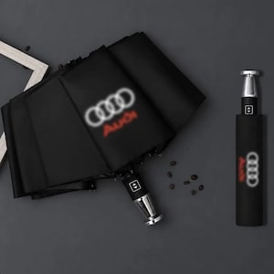 Op de afbeelding: Een zwarte opvouwbare paraplu met het Audi-logo in wit en rood. De paraplu heeft een metalen handvat met een knop en wordt zowel open als dicht getoond. Het ontwerp bevat het embleem met vier ringen en de merknaam Audi in rode tekst.