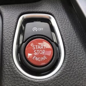 Op de afbeelding: Een ronde rode start-stopknop voor de motor met witte tekst in een zwarte behuizing. Boven de knop bevindt zich een kleiner zwart paneel met een wit A OFF-symbool. Het geheel is omlijst door een zilverkleurige metalen rand.