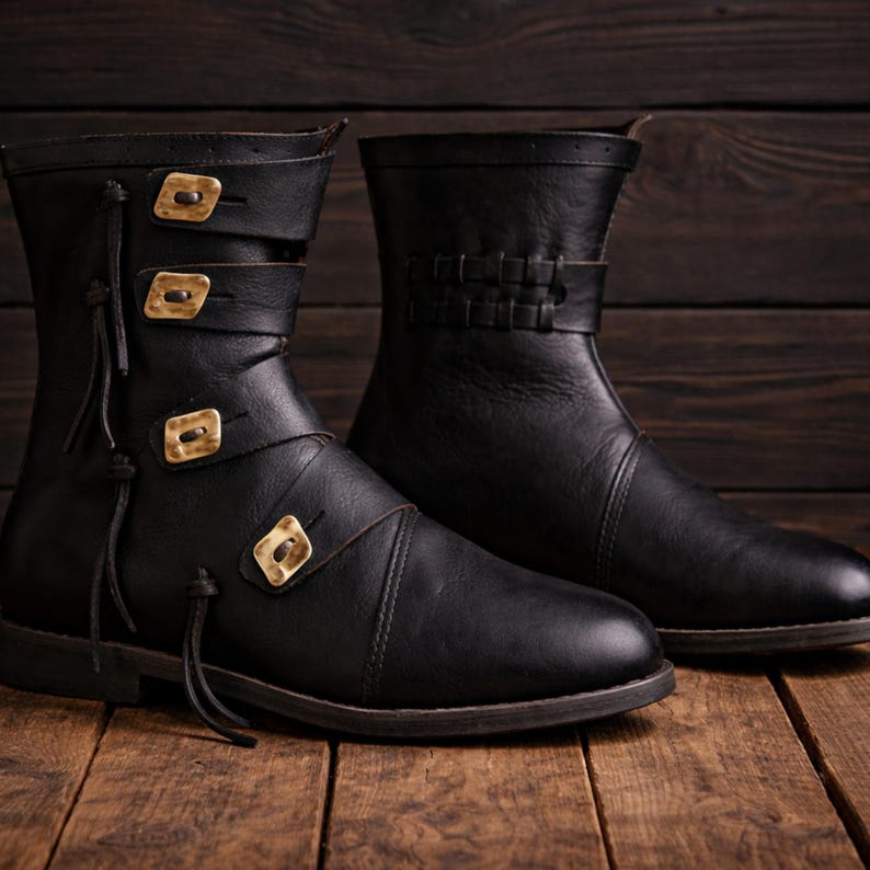 May include: Two black leather boots with decorative gold-colored buckles and straps. One boot features three buckles, while the other has a woven leather detail. The boots are set against a dark wood background.