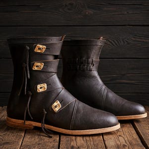 May include: A pair of dark brown leather boots with decorative straps and buckles. The boots have a mid-calf height and a light-colored sole. The buckles are a light tan color. The boots are displayed on a dark wooden surface.