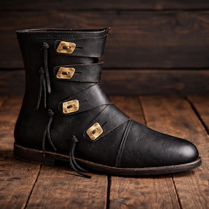 May include: Black leather boot with three straps and decorative gold-colored square buckles. The boot has a rounded toe and a short shaft, with decorative leather ties on the side. The boot is set on a wooden surface.