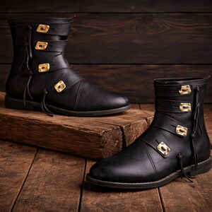 May include: A pair of black leather boots with gold-colored square buckles and straps. The boots have a flat sole and decorative tassels. The boots are displayed on a wooden surface.