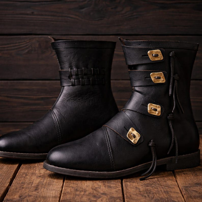 May include: A pair of black leather ankle boots with decorative gold-colored buckles and straps. One boot features a woven leather strap, while the other has three buckles. The boots are displayed on a wooden surface.