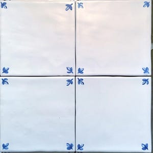 May include: Four white ceramic tiles arranged in a square, each with a blue floral corner design. The tiles are square and have a glossy finish, with the blue floral accents adding a touch of color and detail to the otherwise plain white surface.