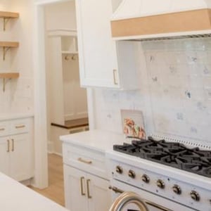 May include: A kitchen interior featuring white cabinetry with gold hardware, a white range stove with black burners, a white range hood with a wood accent, and open wooden shelving. A tiled backsplash with blue patterns is visible behind the stove.