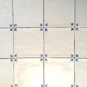 May include: A grid of square off-white ceramic tiles featuring a repeating blue leaf pattern at each corner. When four tiles meet, the corner motifs combine to form a small blue floral accent at the intersection of the grout lines.