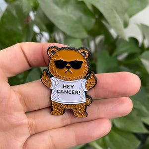 May include: A brown bear enamel pin wearing sunglasses and a white t-shirt with the text "HEY CANCER!" The bear is giving the middle finger with both hands. The pin has a black outline.