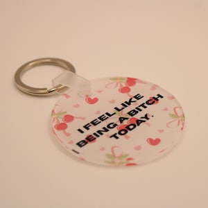 May include: A clear, round keychain with a silver key ring. The keychain has a white background with a pattern of pink cherries, hearts, and green leaves. The text "I FEEL LIKE BEING A BITCH TODAY." is printed in black.